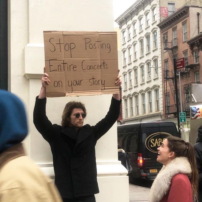 This meme features a man holding a cardboard sign that reads "Stop posting entire concerts on your story" in a sarcastic tone. The image is likely a humorous commentary on social media users who share lengthy, unedited recordings of live events on their social media stories. The sign is held by a man in a humorous, exaggerated pose, adding to the comedic effect.